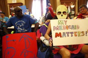 An office is filled with members of ADAPT - two at the forefront have signs that read My Medicaid Matters and ADAPT. One of the protesters is wearing a skull mask