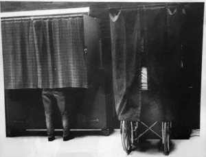 A black and white photo of two voting booths with curtains. Behind one curtain is a person in a wheelchair and behind the other voting booth curtain is someone standing. 