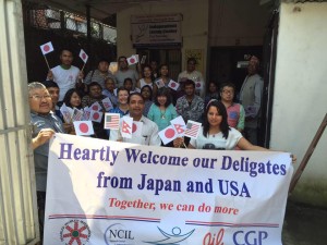 Nepalese Delegation of 23 people hold American and Japanese and Neopalese flags and a banner that reads Heartly Welcome our Deligates from Japan and USA - Together we can do more