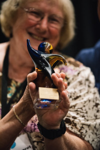 Two hands clasp the Women's Caucus Award, which is multi-colored glass shaped like and abstract person. Women's caucus Co-Chair Mary Margaret Moore smiles in the background while presenting the award. 