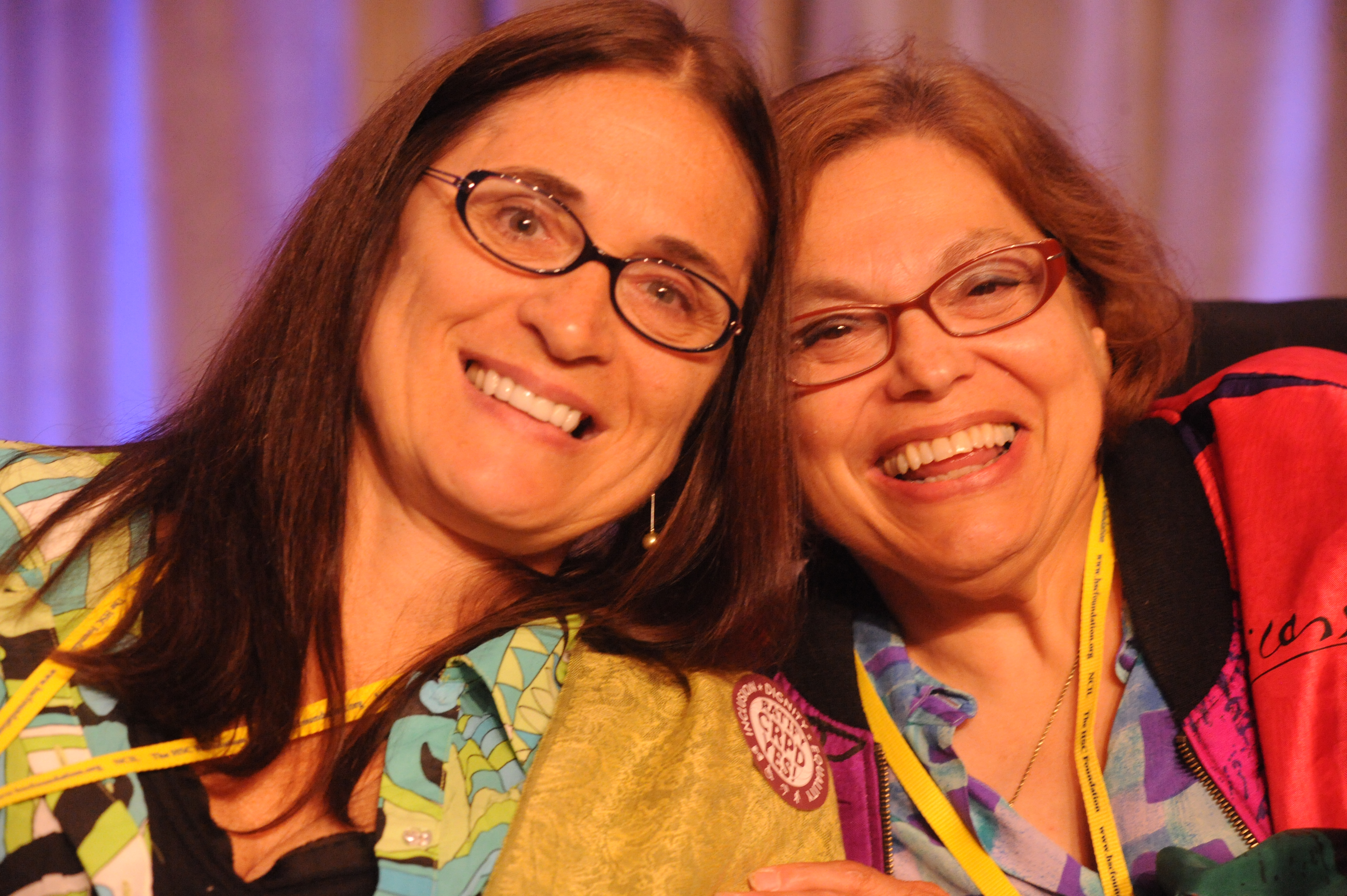 Marca Bristo and Judy Heumann clasp hands and smile for a photo from the stage at the 2007 NCIL Annual Conference on Independent Living