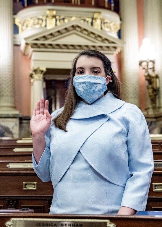 Representative Jessica Benham, a young woman with brown hair wearing a blue face mask and blue coat, stands at a desk and raises her right hand as she takes the oath of office.