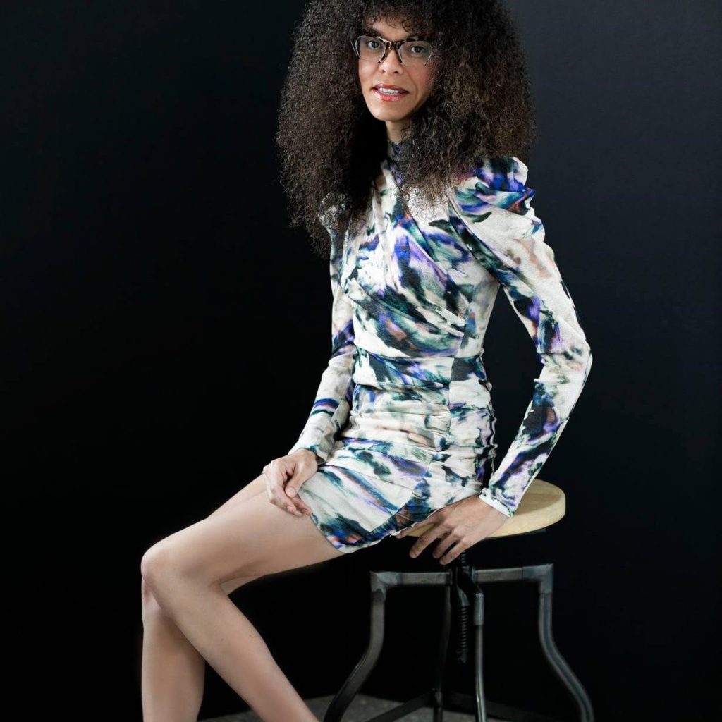 Reyma McCoy McDeid, a tall, light-skinned Black woman with long curly hair and glasses leaning on a tall stool
