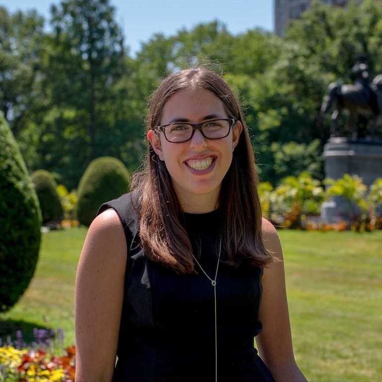 Image: Jessica, a white woman with long brown hair and glasses, is standing in the Boston Common, smiling. 