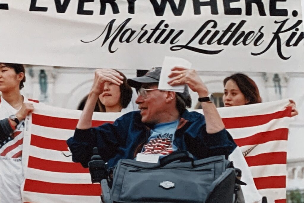 Paul Spooner at a NCIL Rally in the 1990s. He is speaking from the stage in front of the NCIL banner and a flag held by three Japanese people.