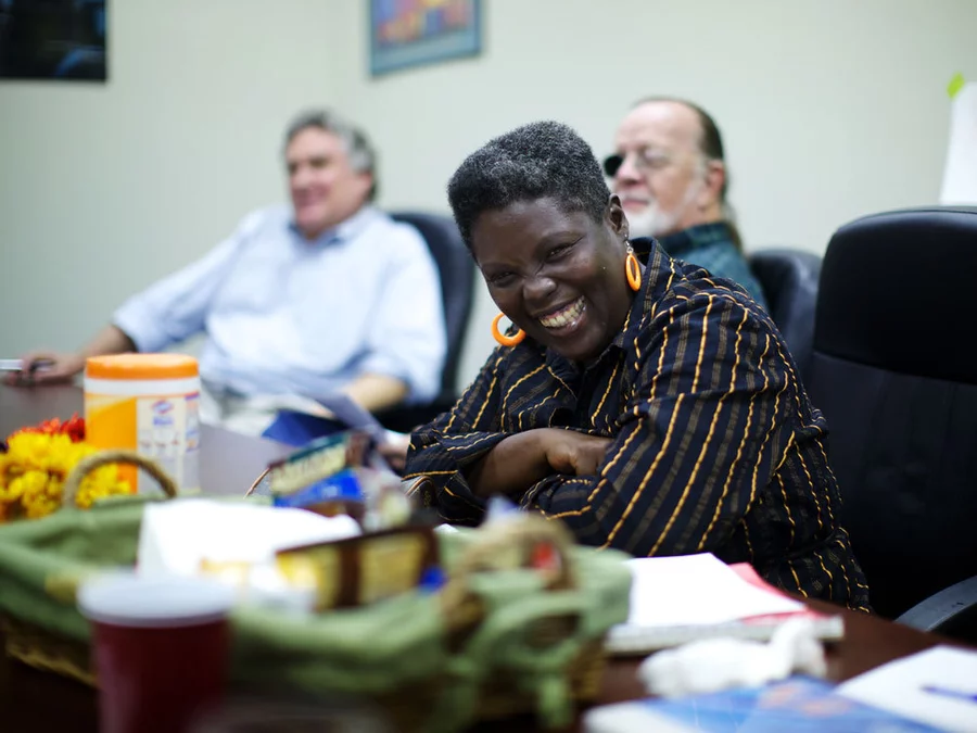 Photo of Lois Curtis, a Black woman with dark skin and short Afro smiling joyfully as she sits in a room among friends. Photo courtesy of NPR.