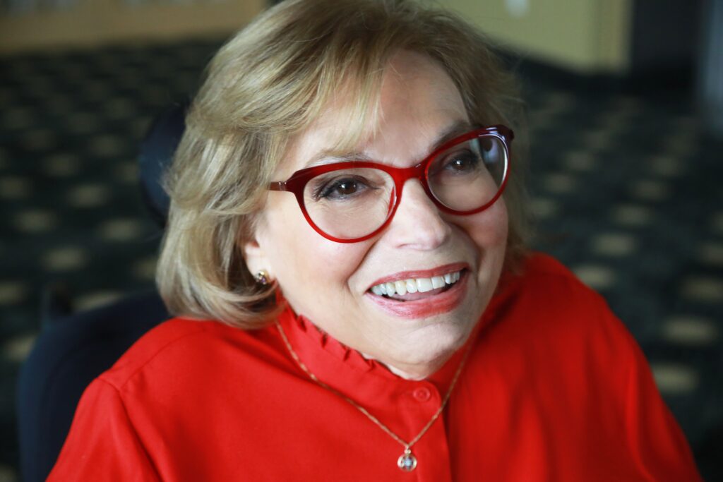 A headshot of Judy Heumann, smiling warmly and looking up toward the camera. She is wearing a bright red shirt and bright red glasses and lipstick.