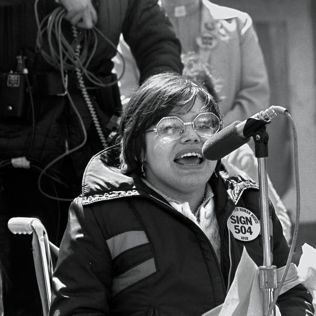 A black and white photo of a young Judy Heumann as she speaks into a microphone. She is wearing a winter coat with a “Sign 504” sticker. 
