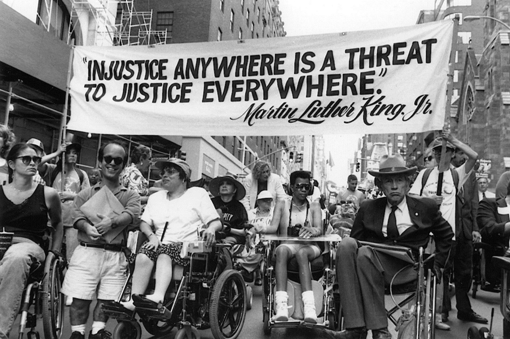 Judy Heumann, Marca Bristo, Justin Dart, and many others lead the 1993 NCIL March to the Capitol. A large banner says “Injustice Anywhere is a Threat to Justice Everywhere – Martin Luther King, Jr.”