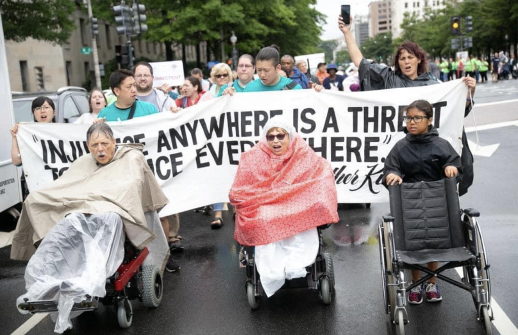 Image: Judy Heumann, Kelly Buckland, and many others lead the 2018 NCIL March to the Capitol. They are wearing ponchos and leading chants while marching in the pouring rain. Anna L-T is pushing Justin Dart’s wheelchair next to them.