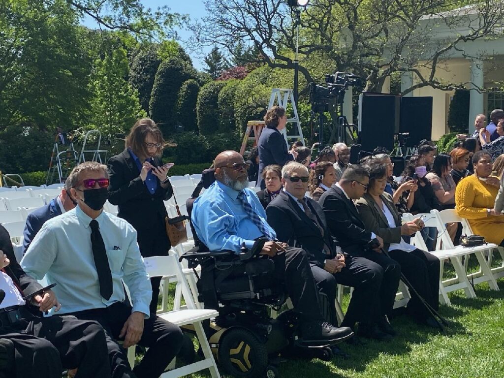 NCIL Executive Director Theo Braddy, a Black man with a salt and pepper beard and a power wheelchair sits amongst guests at the outdoor signing ceremony. 